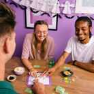 Three friends playing a board game together with a purple wall and Halloween decorations in the background.
