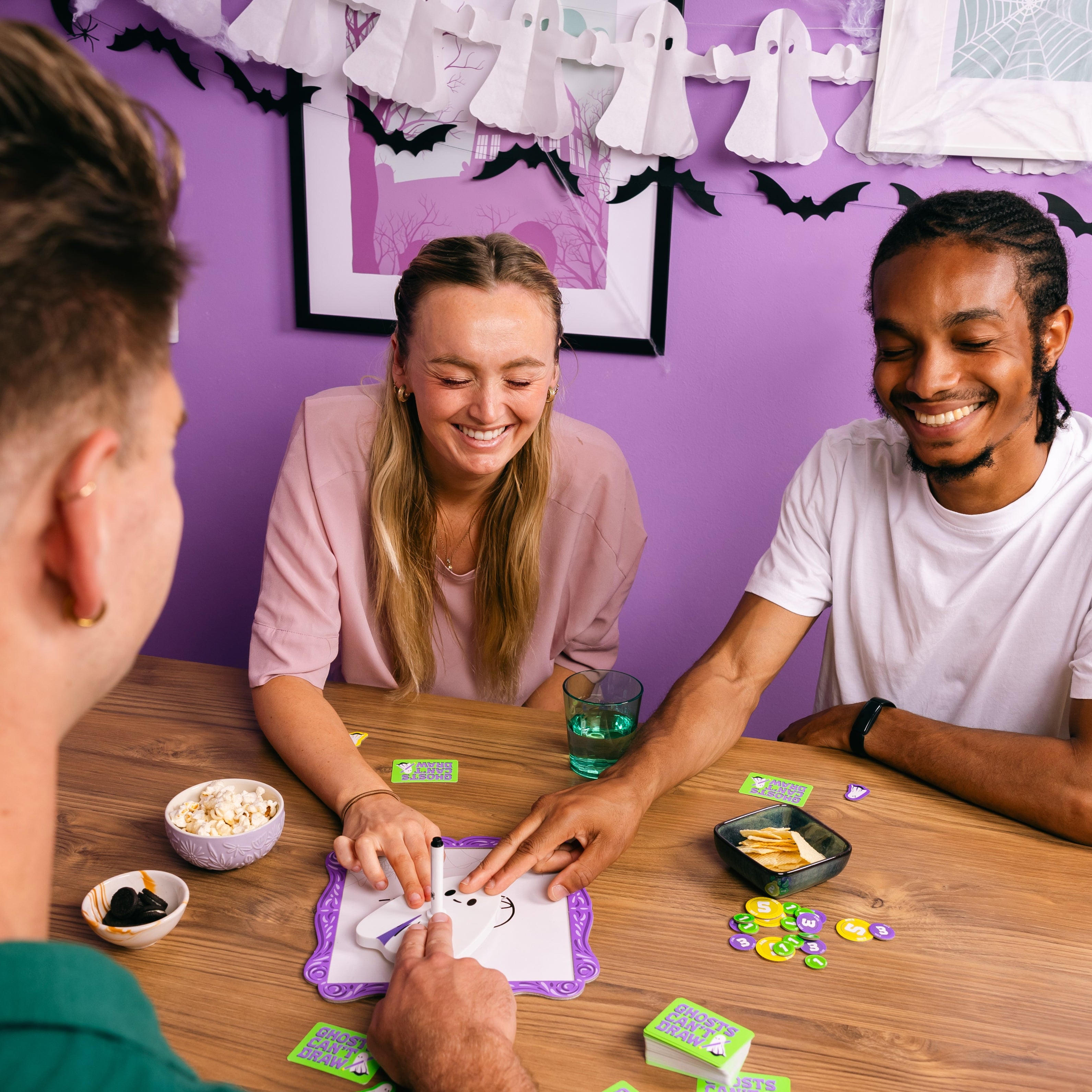 Three friends playing a board game together with a purple wall and Halloween decorations in the background.